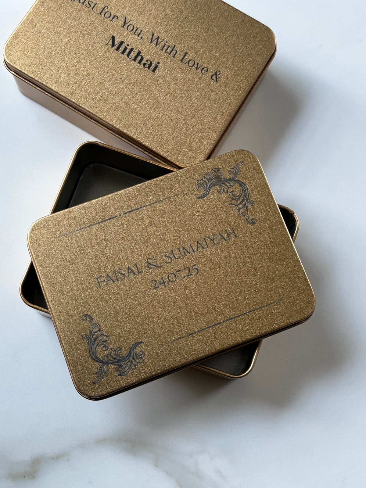 Two personalized gold metal gift boxes with matching lids, shown on a light marble surface. One box is open, revealing a dark interior, and the other lid is placed above it.