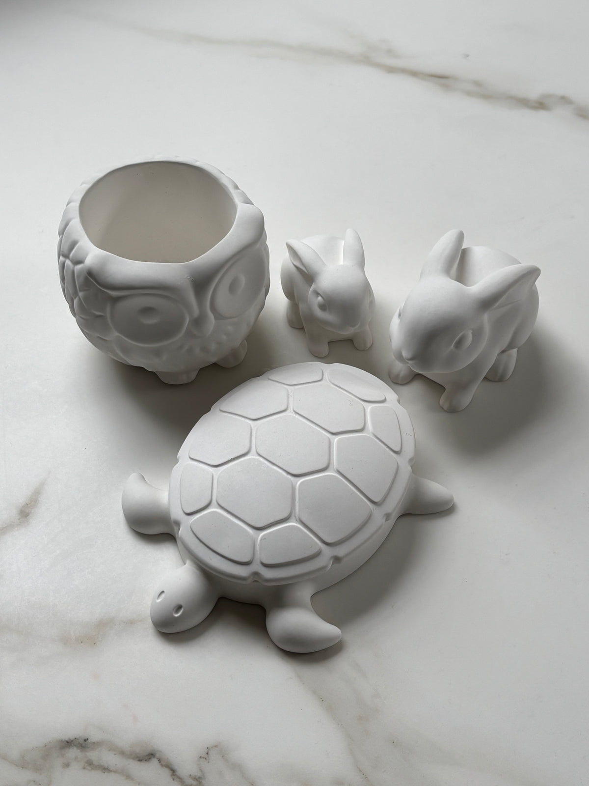 A set of four white ceramic animal-shaped containers, including an owl, a turtle, and two rabbits, arranged on a white marble surface.